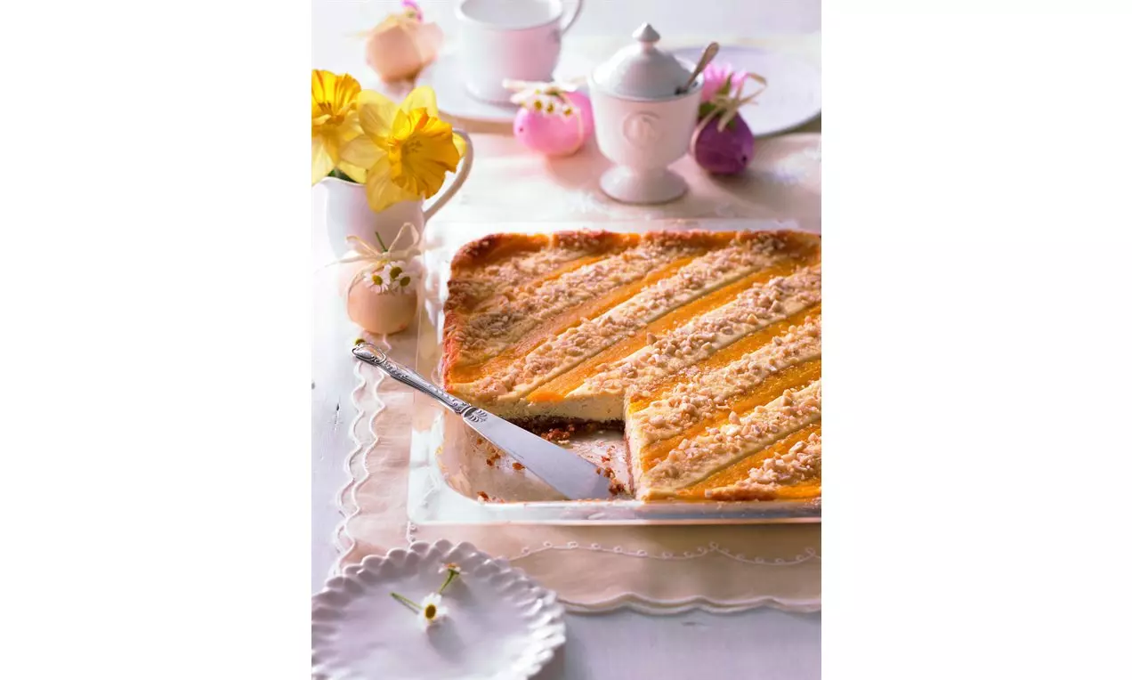 Prăjitură dreptunghiulară cu straturi aurii de mango și nuci mărunțite pe o tavă, alături de flori galbene și porțelan alb