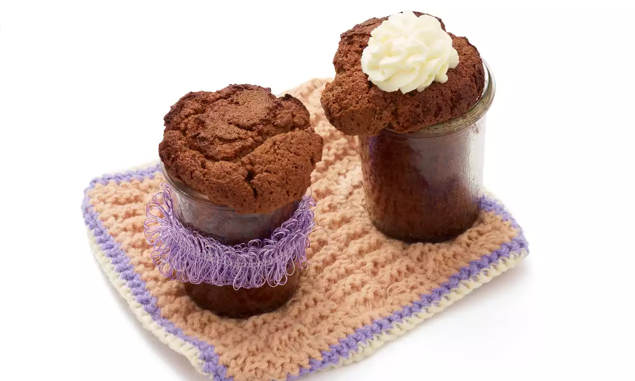 Zwei gläserne Schokoladenküchlein mit luftig gebackener, dunkelbrauner Oberfläche, eines mit Sahnehäubchen auf gehäkelter Unterlage drapiert