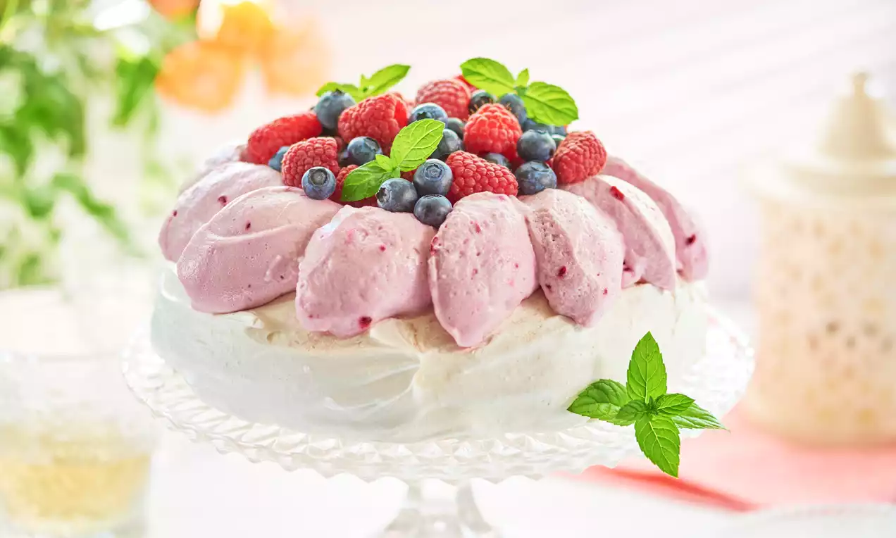 Beza z chrupiącą skorupką i puszystym różowym kremem, udekorowana malinami, borówkami i listkami mięty na szklanym talerzu