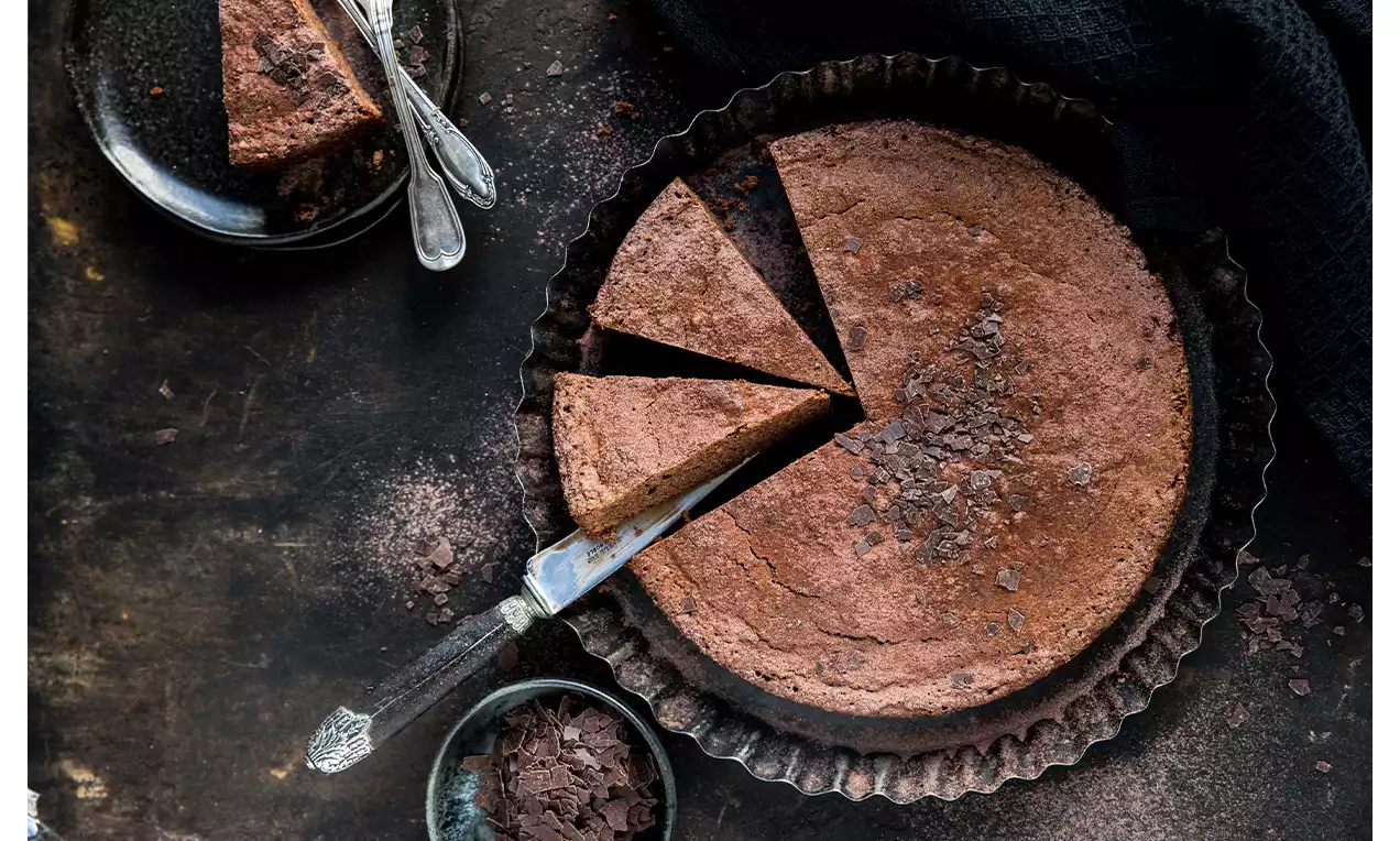 Runder Haselnuss-Schoko-Kuchen in geriffelter Backform, angeschnitten mit Schokoladenstücken und Krümeln, daneben Teller mit Kuchenscheibe