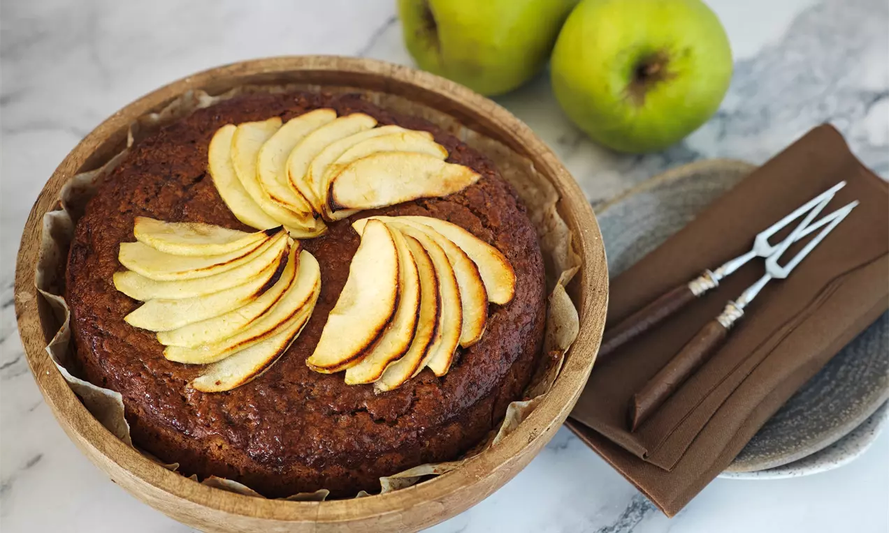 Златисто-кафяв ябълков сладкиш в кръгла форма с карамелизиран повърхностен слой, украса от тънки резени ябълки и два зелени плода на заден план