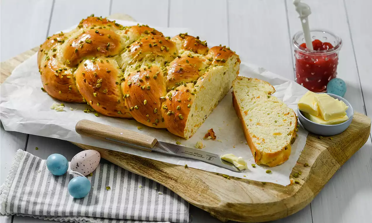 Tresse briochée dorée à la croûte brillante parsemée de pistaches, tranchée sur du papier cuisson avec du beurre et confiture à côté