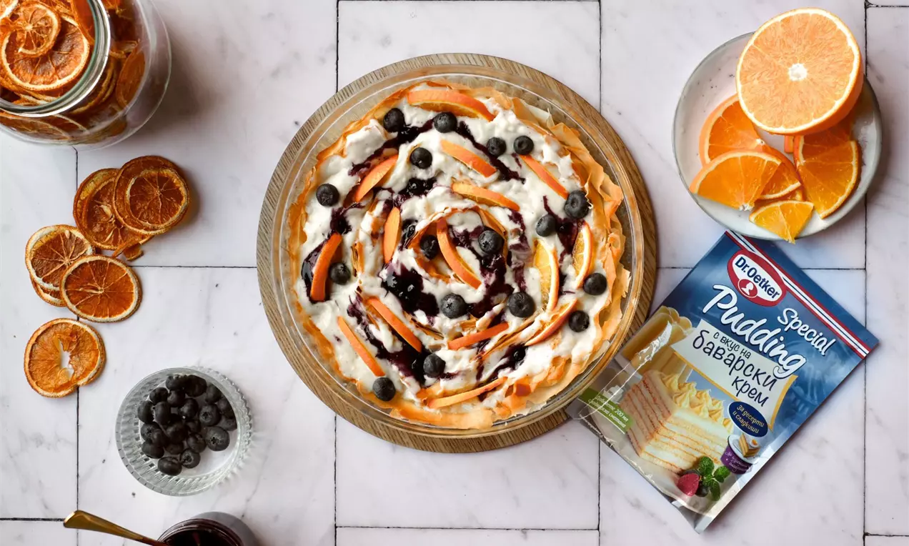 Кръгла крем пита с жълтеникаво хрупкава основа, пухкав бял крем, парченца портокал и свежи боровинки, сервирана върху бяла плоча