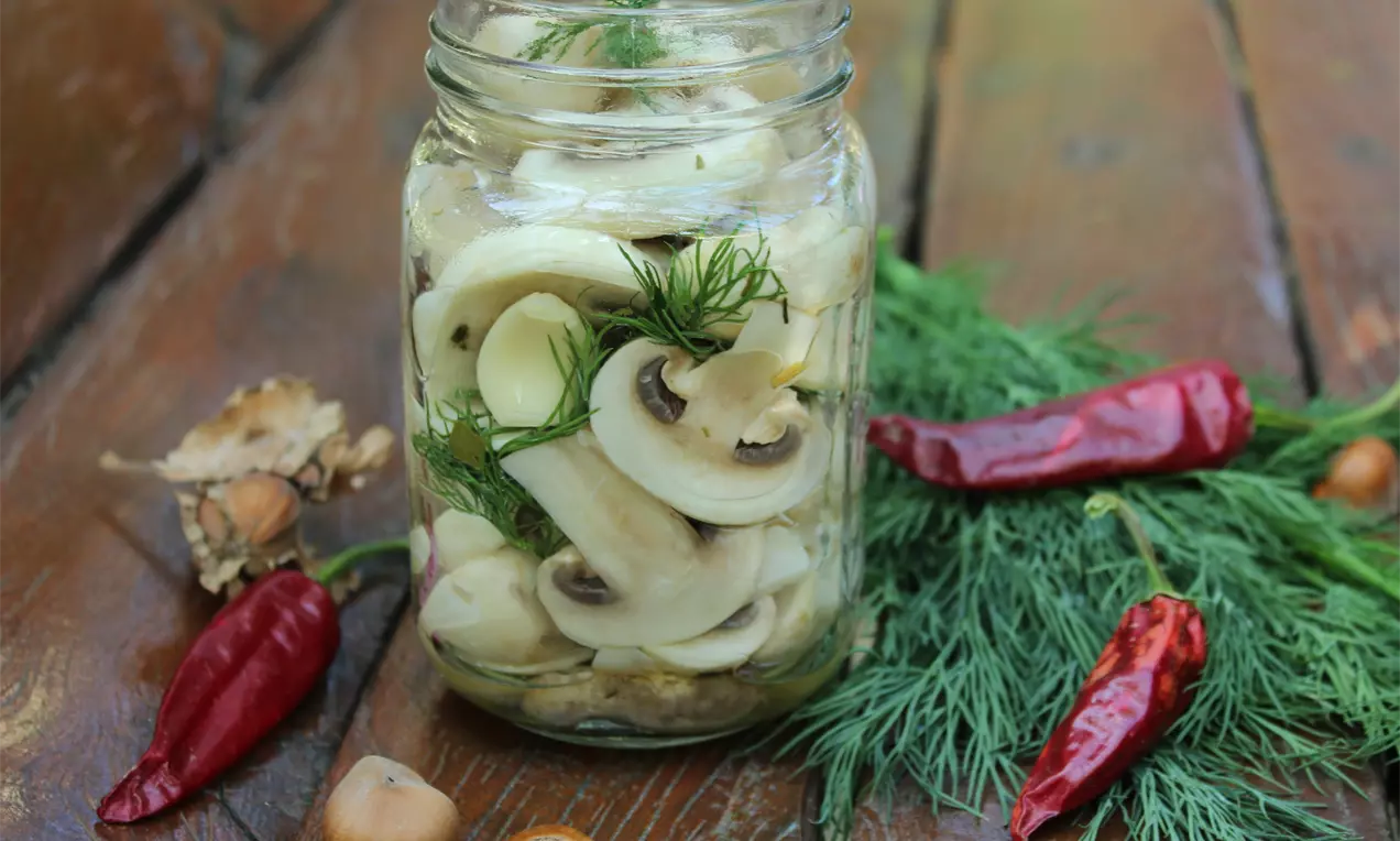 Светли резени гъби с нежна текстура, подредени в стъклен буркан с украса от свеж копър, заобиколени от люти чушки и орехи върху дървена маса
