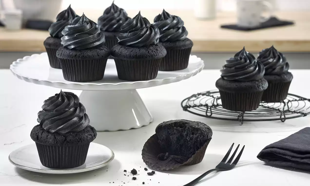 Schwarze Cupcakes mit glänzender, spiralförmig aufgetragener Creme auf weißer Tortenplatte, ein angeschnittener Cupcake auf Teller mit Gabel