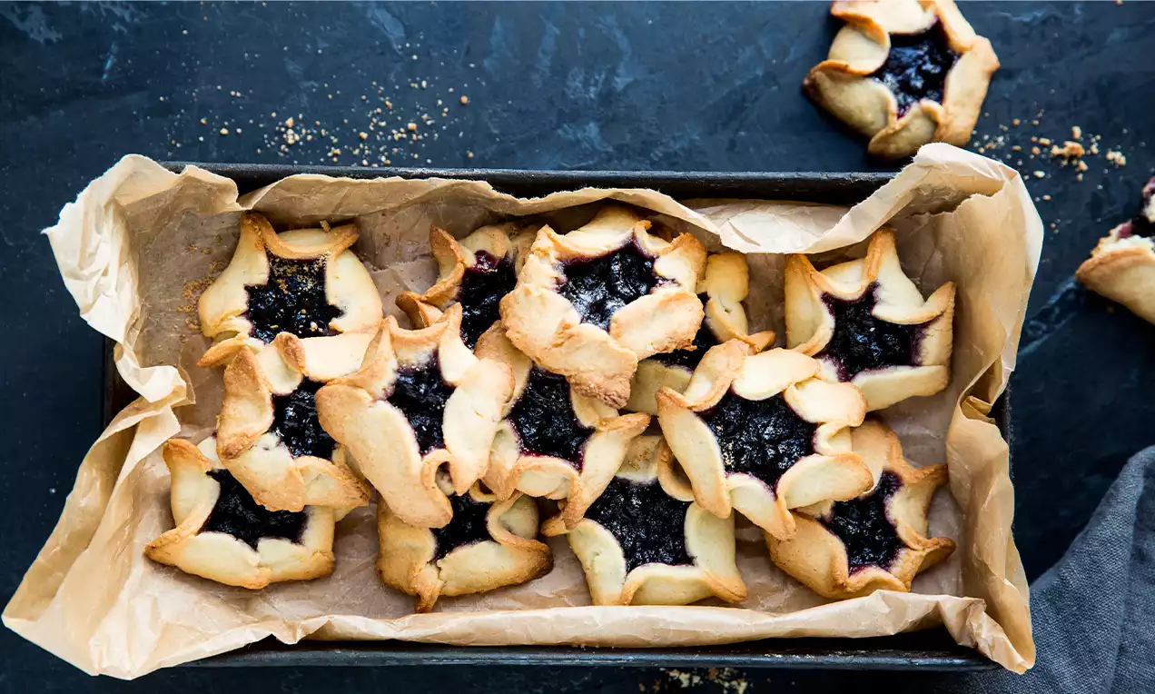 Biscuits dorés en forme d’étoiles garnis de confiture foncée sur papier cuisson fripé dans un plateau noir