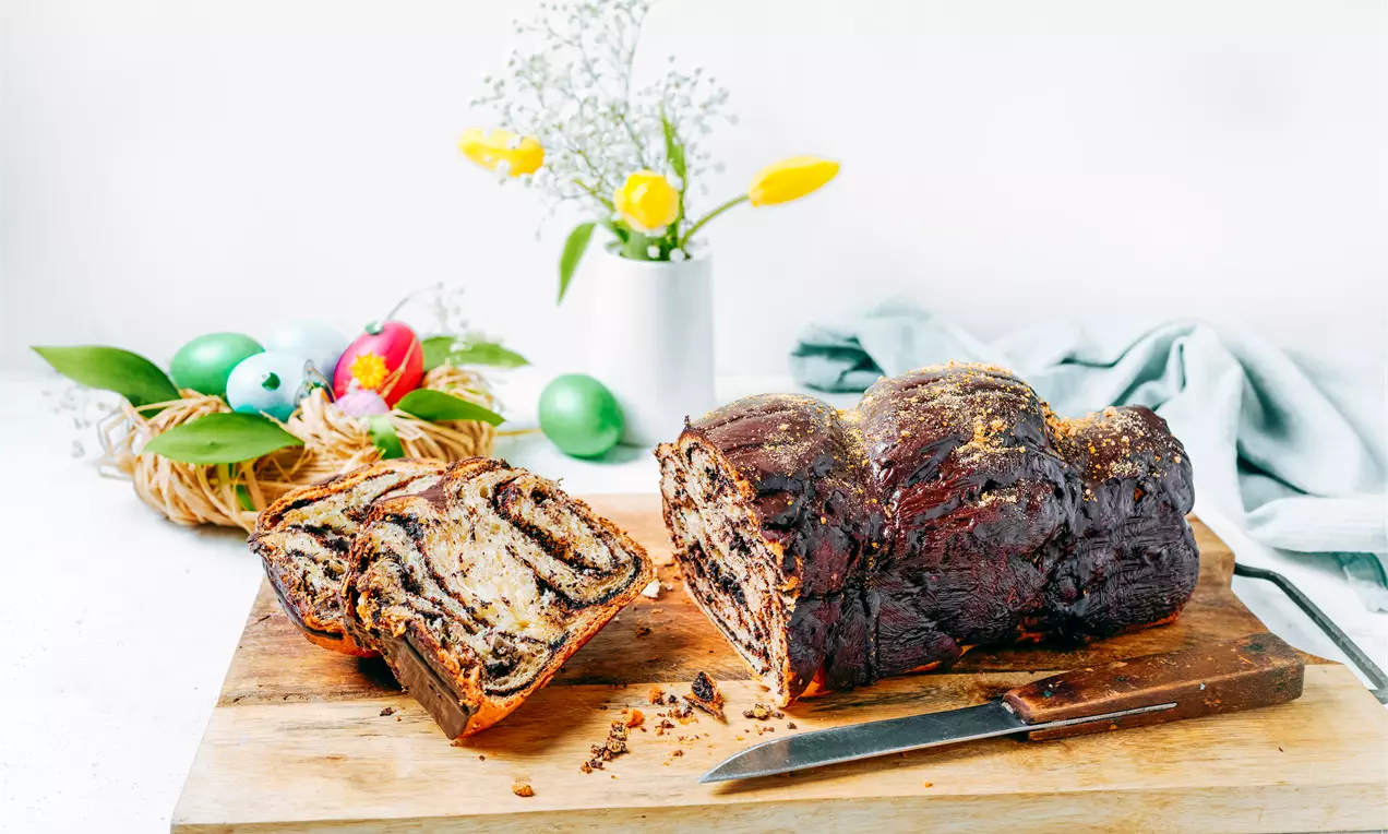 Козунак с тъмнокафява глазура върху златиста коричка, с разрез, разкриващ спирали от шоколадово тесто, върху дървена дъска с нож