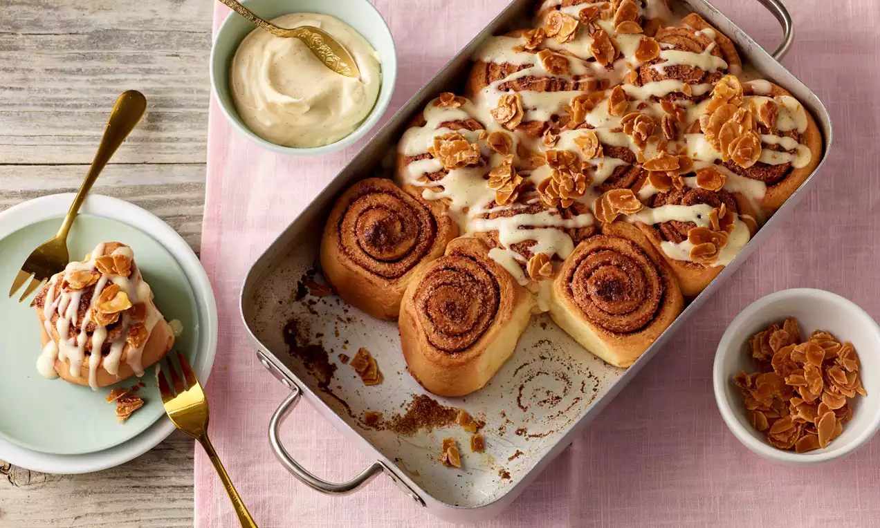 Goldbraune Zimtschnecken dicht aneinandergereiht in einer silbernen Auflaufform, mit Zuckerguss und gerösteten Mandelblättchen verziert