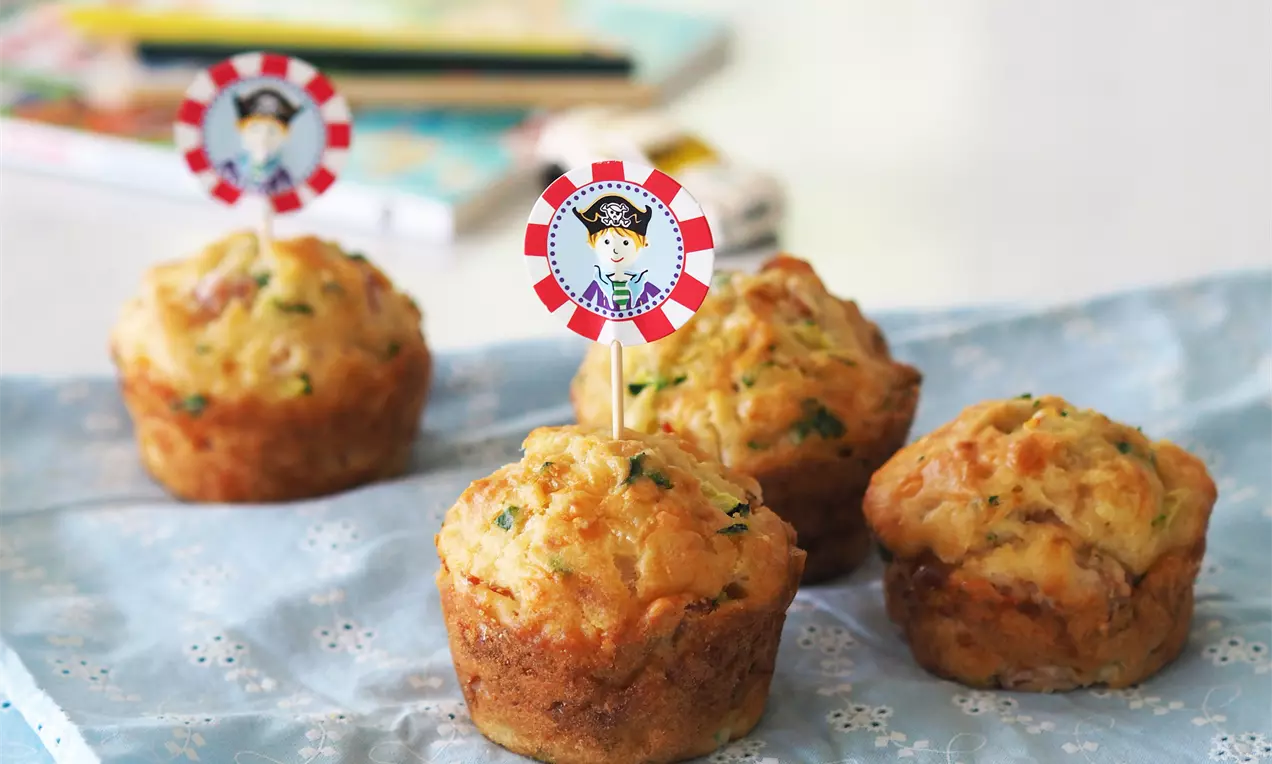 Златисто-кафяви мъфини с поръски зелени подправки, подредени върху синя покривка, украсени с пиратски флагчета на дървени клечки