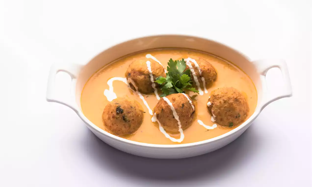 Golden-brown round koftas in silky orange curry garnished with white cream drizzles and fresh green coriander in a white serving dish