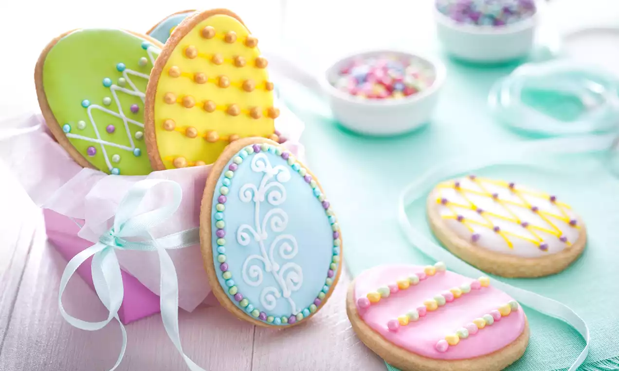 Påskkakor formade som färgglada ägg, dekorerade med glasyr och pärlor, ligger på bord och i rosa ask med vitt silkespapper och blå snöre
