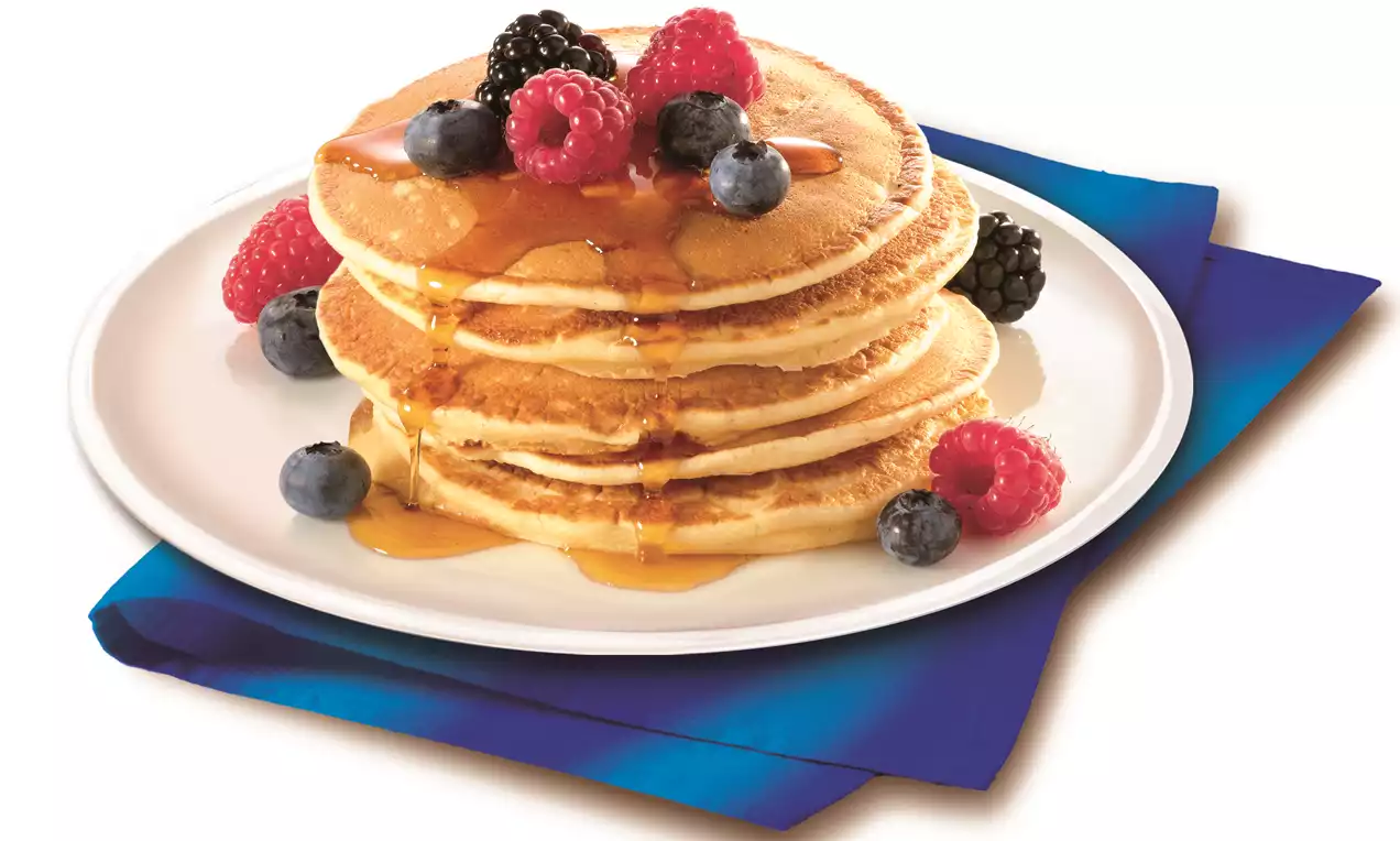 Een stapel goudbruine pannenkoeken overgoten met glanzende siroop, gegarneerd met frambozen, bosbessen en bramen op een wit bord op een blauwe ondergrond