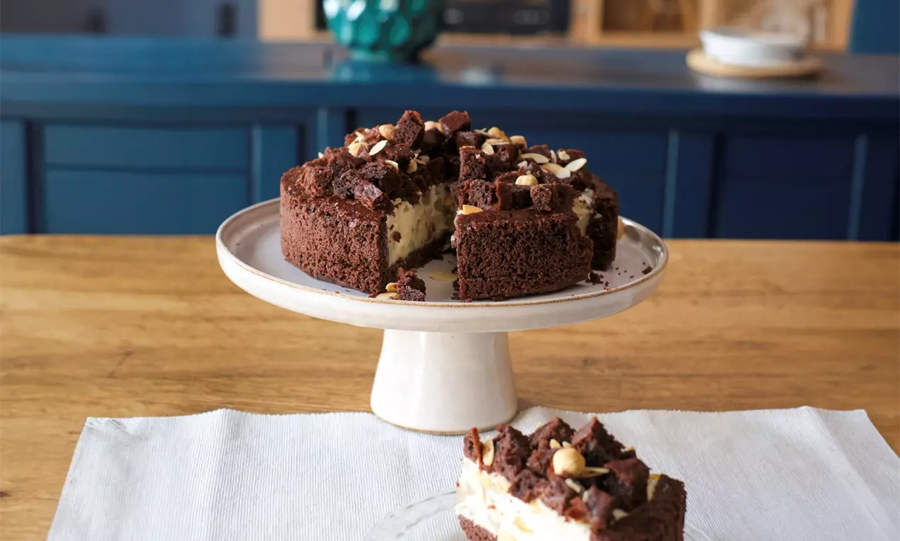 Торта с баварски крем с лека текстура, поширани круши и хрупкави парченца бадеми върху плътен тъмнокафяв блат, сервирана на порцеланова стойка