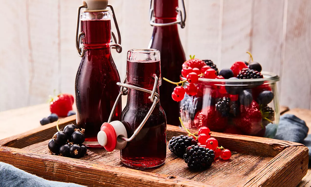 Тъмночервен сироп в стъклени бутилки със закопчалки, заобиколени от свежи горски плодове в буркан и върху дървена табла