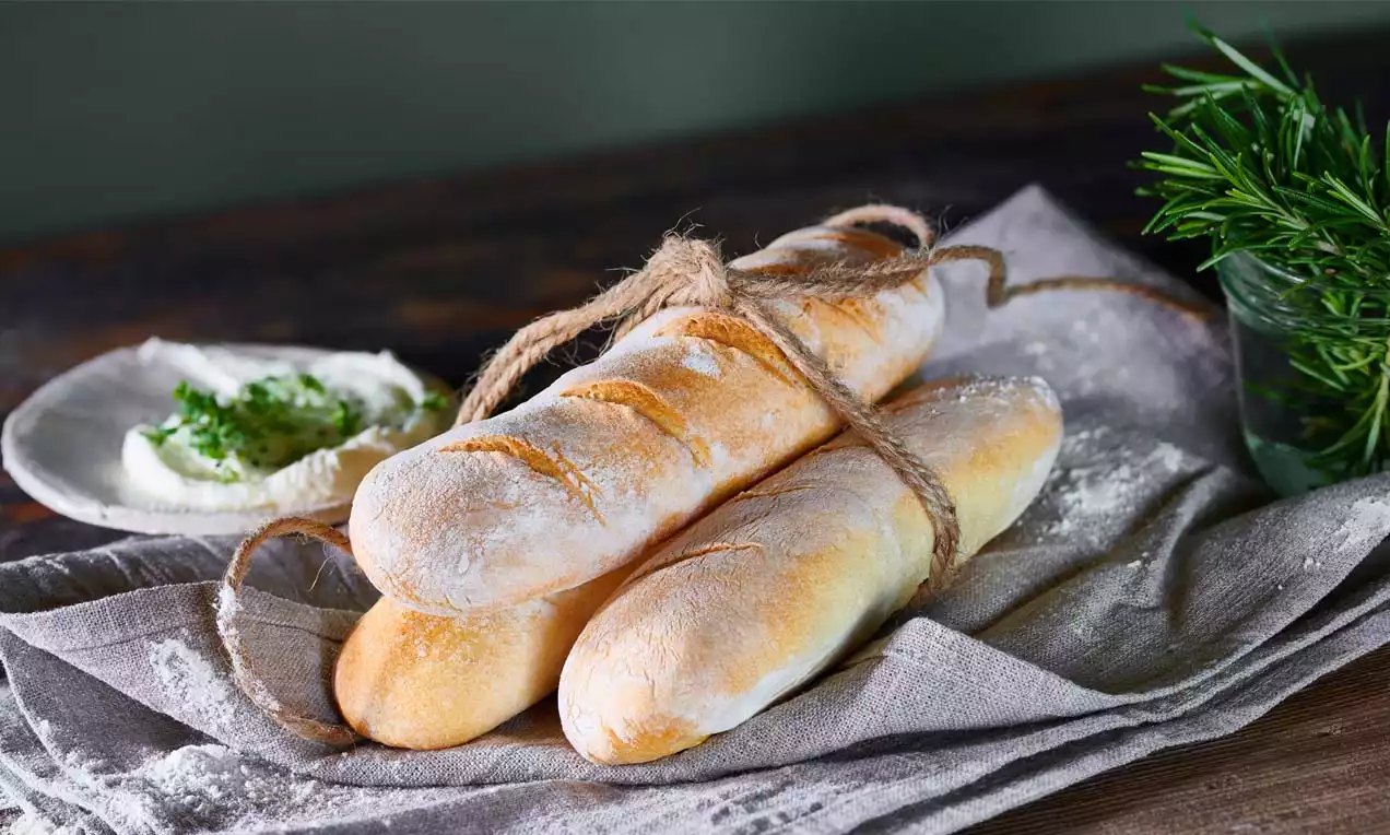 Drei goldgelbe Mini-Baguettes mit feiner Mehlschicht, zusammengebunden mit rustikalem Seil auf grauem Stofftuch, daneben Kräuterquark und Rosmarinzweig