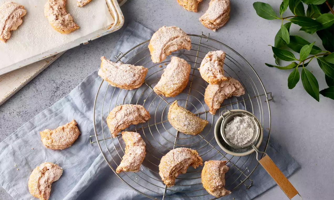 Burgenländer Kipferln hellbraun und zart, leicht gebogene Form mit Puderzucker bestäubt auf einem runden Rost neben einem Sieb mit Zucker