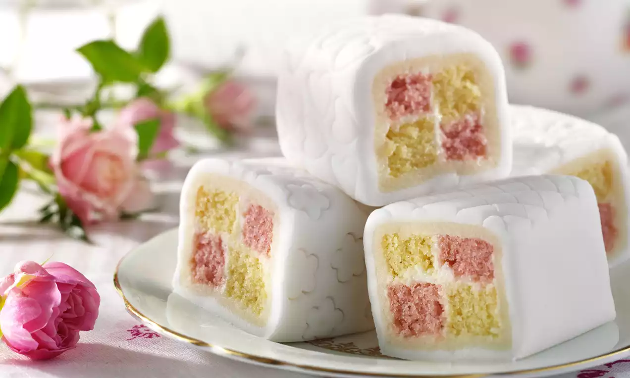 Wit omhulde cakeblokjes met roze en gele vierkante laagjes, versierd met een patroon van marsepein, gepresenteerd op een wit bord met bloemen