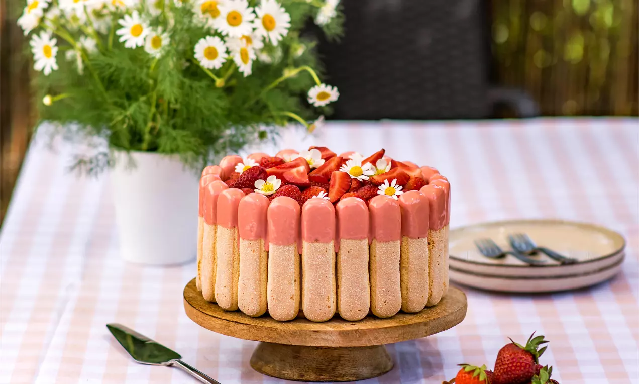Торта Шарлота с високи бишкоти, покрита с розов мус, свежи ягоди и цветчета, поставена върху дървена стойка на каре маса