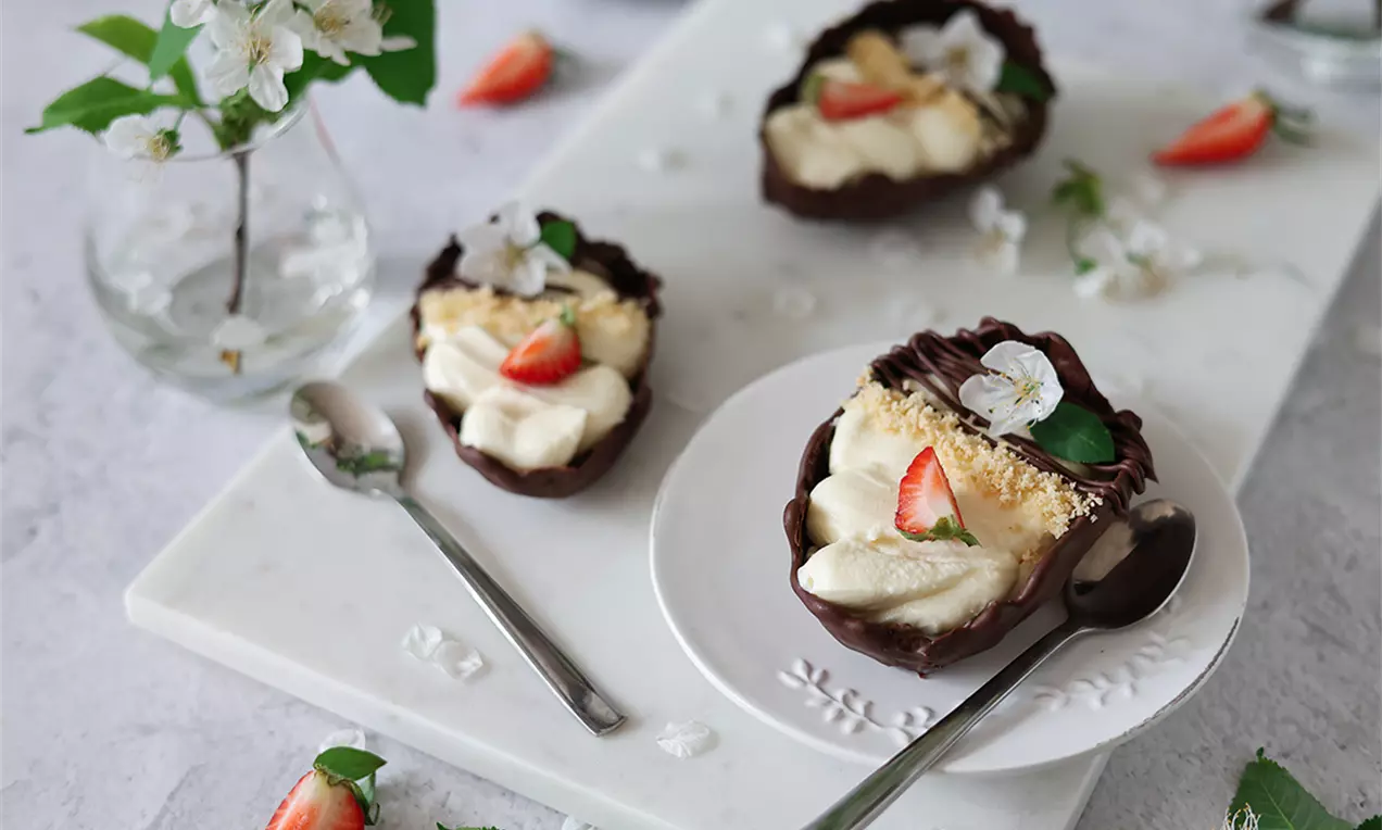Половинки тъмни шоколадови яйца, пълни с кремообразен ванилов чийзкейк, украсени с пресни ягоди и цветчета върху бяла чиния