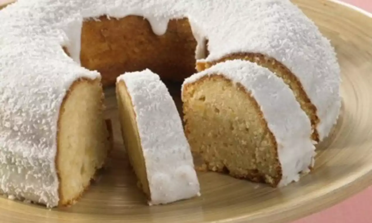 Round coconut cake with a hole in the center, covered in white icing and desiccated coconut, sliced on a wooden plate with a pink backdrop