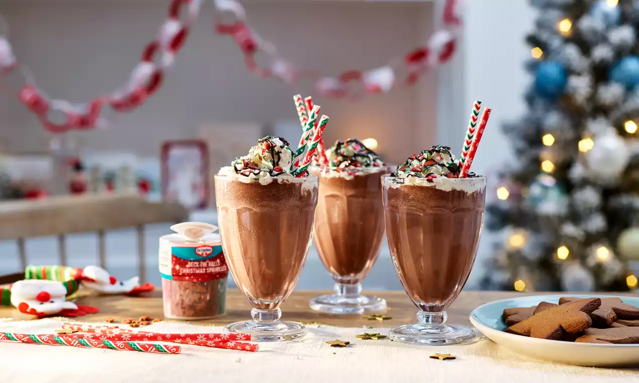 Three tall glasses of rich chocolate milkshake topped with whipped cream, sprinkles, and striped straws on a festive table
