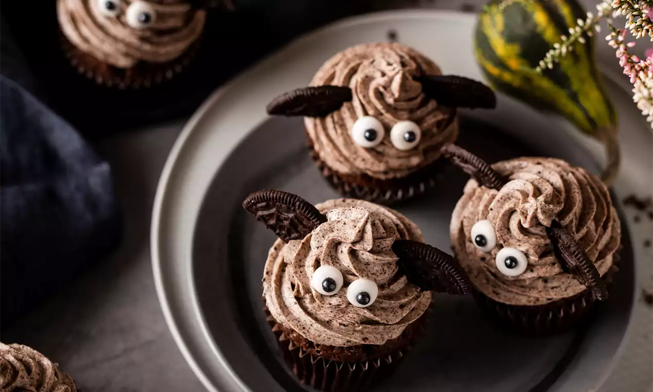 Helovyno keksiukai su šokoladinės spalvos kremu, garbanotais sūkurių raštais, dekoruoti akimis ir sausainių sparneliais pilkoje lėkštėje