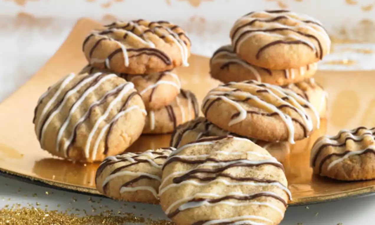 Vanillekleurige zandkoekjes met een zachte textuur, gedecoreerd met witte en bruine chocolade drizzles, gerangschikt op een glanzende goudkleurige schaal