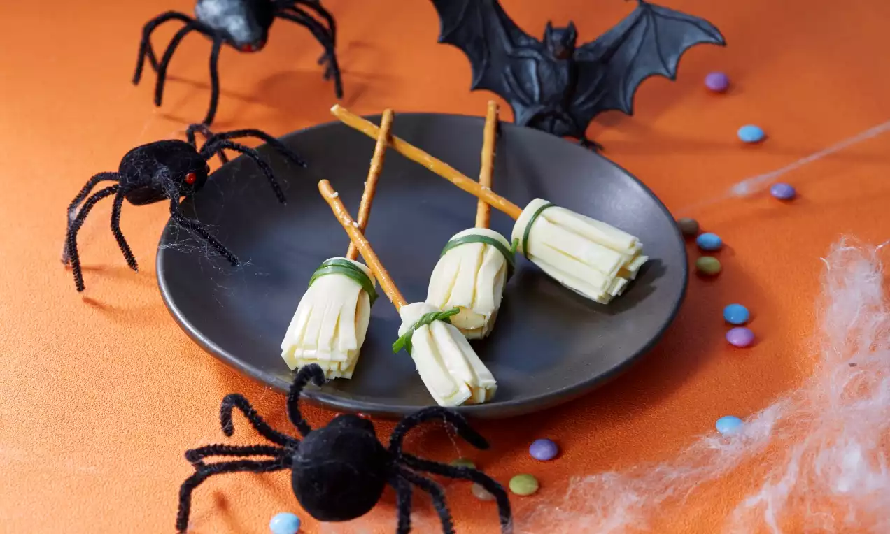 Balais en bretzel doré et fromage blanc frangé noués de ciboulette sur une assiette noire entourée d’araignées et d’une chauve-souris