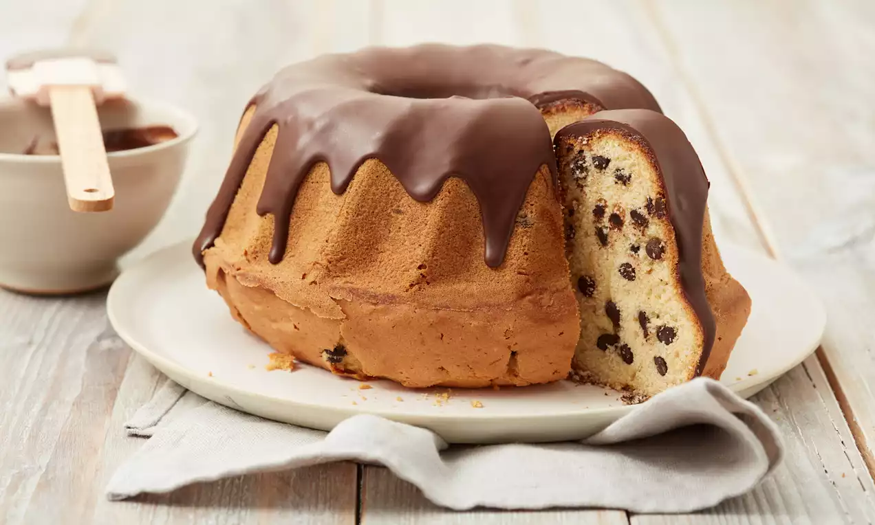Runder Gugelhupf mit goldbraunem Teig, dunklen Schokostückchen und glänzendem Schokoladenguss auf einem hellen Teller mit Stoffserviette