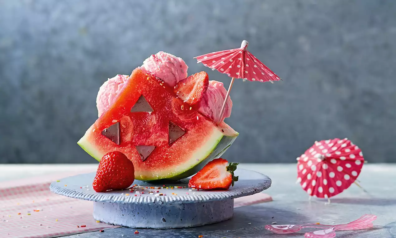 Ein Stück rote Wassermelone mit Dreieck-Ausschnitten, rosa Eis und Erdbeeren garniert, dekoriert mit einem gepunkteten roten Papier-Schirm