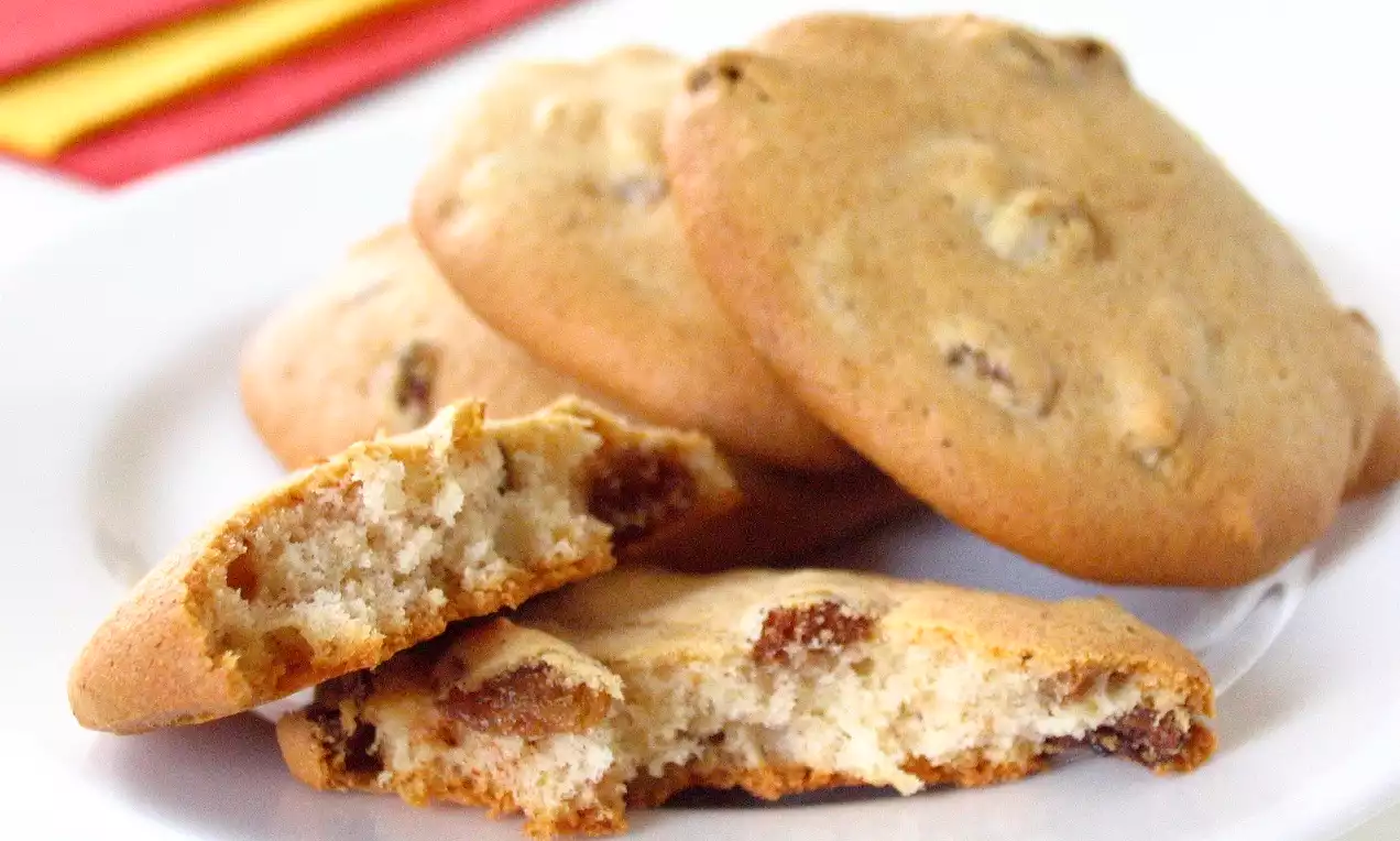 Yuvarlak şekilli, altın kahverengi, yumuşak dokulu üzümlü kurabiyeler beyaz bir tabakta bir araya getirilmiş, fon renkli bir peçete ile tamamlanmış