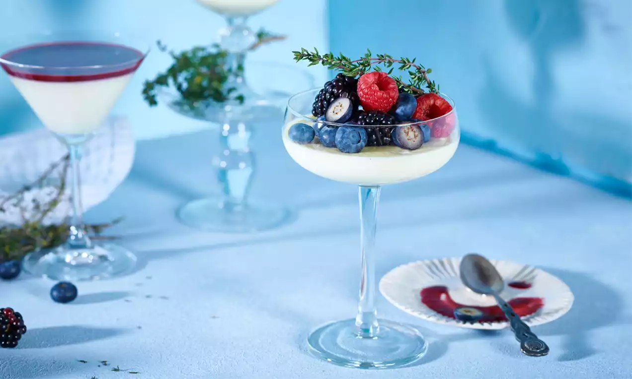 Panna cotta svijetle krem boje u prozirnoj čaši na stalku, ukrašena svježim bobičastim voćem i grančicom timijana na plavoj podlozi