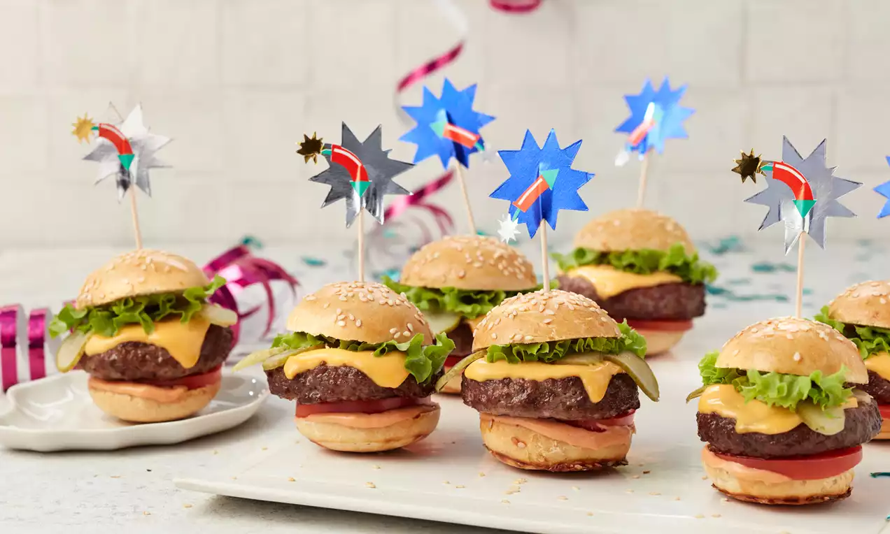 Mini-Burger mit goldbraunem Sesambrötchen, geschmolzenem Käse, grünem Salat und Tomatenscheibe dekorativ auf weißer Platte angerichtet