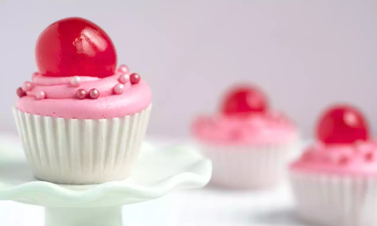 Cupcake met lichtroze glazuur, gegarneerd met een glanzende rode gelatinebol en roze parels op een wit taartplateau met zachte achtergrond