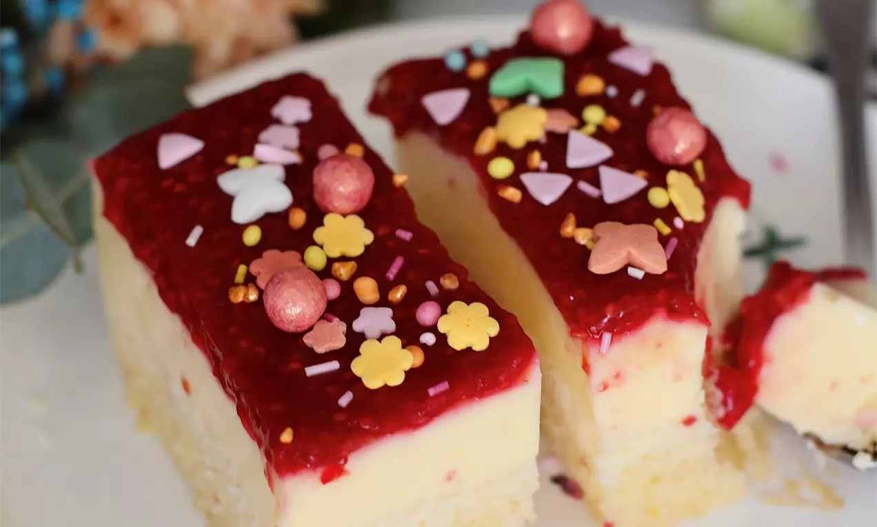 Dvije kremaste kocke na biskvitnom sloju sa sjajnim crvenim malina prelivom, posute šarenim perlicama i šećernim dekoracijama na bijelom tanjiru