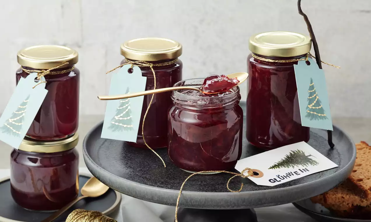 Glühwein-Marmelade in gläsernen Schraubdeckelgläsern mit goldenen Deckeln, tiefrote glänzende Farbe, dekoriert mit hellblauen Geschenkanhängern
