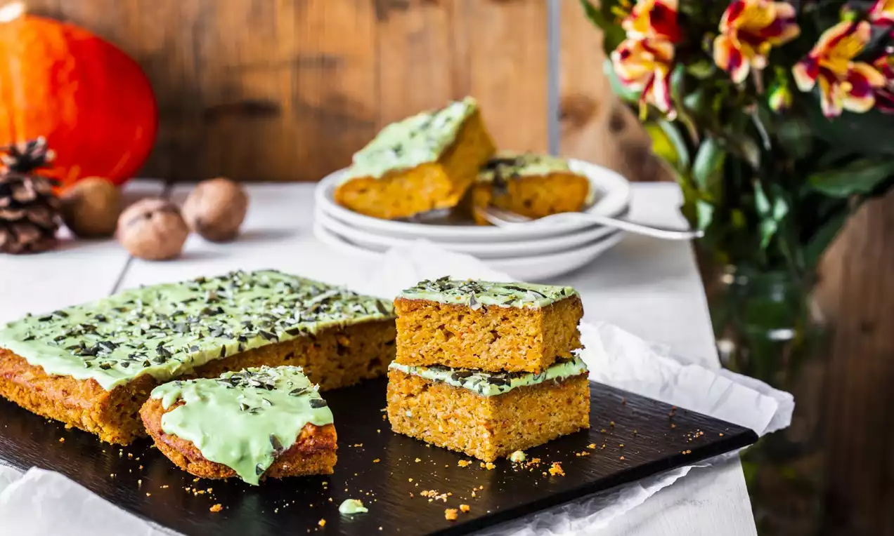 Carrés moelleux de gâteau au potiron orange vif avec un glaçage vert pâle parsemé de graines, disposés sur une planche noire