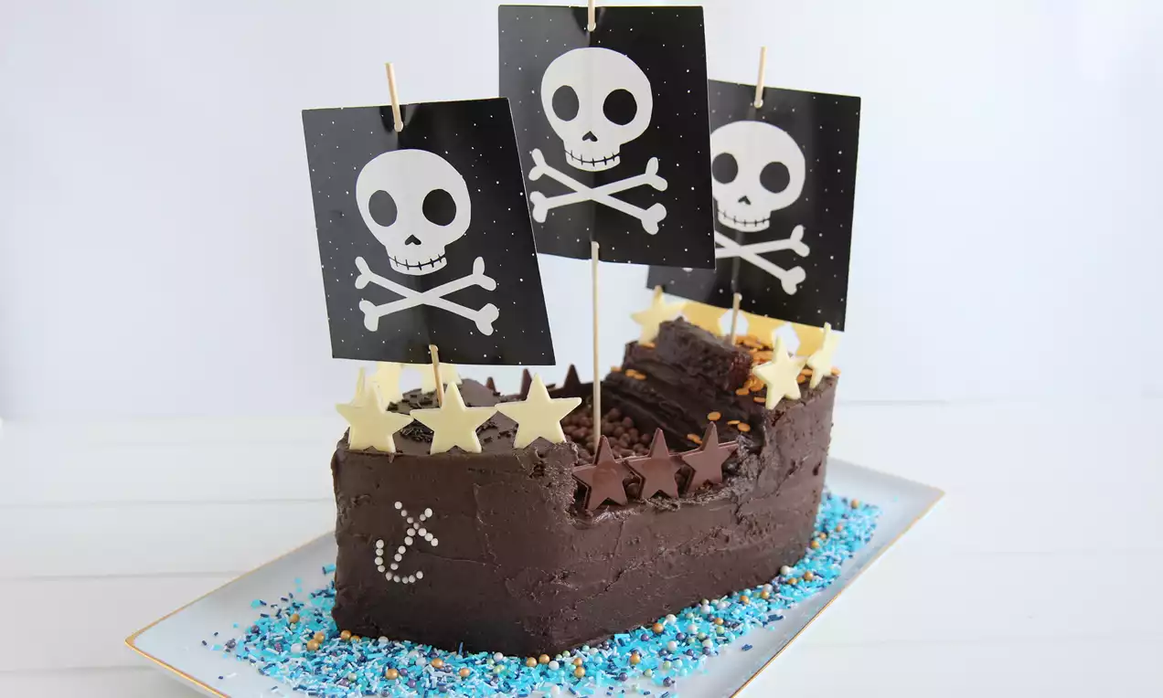 Sjørøverskuteformet sjokoladekake med brunt kremtrekk, svarte seil med hodeskallemotiv, stjernesjokolade og blått sukkerpynt på et fat