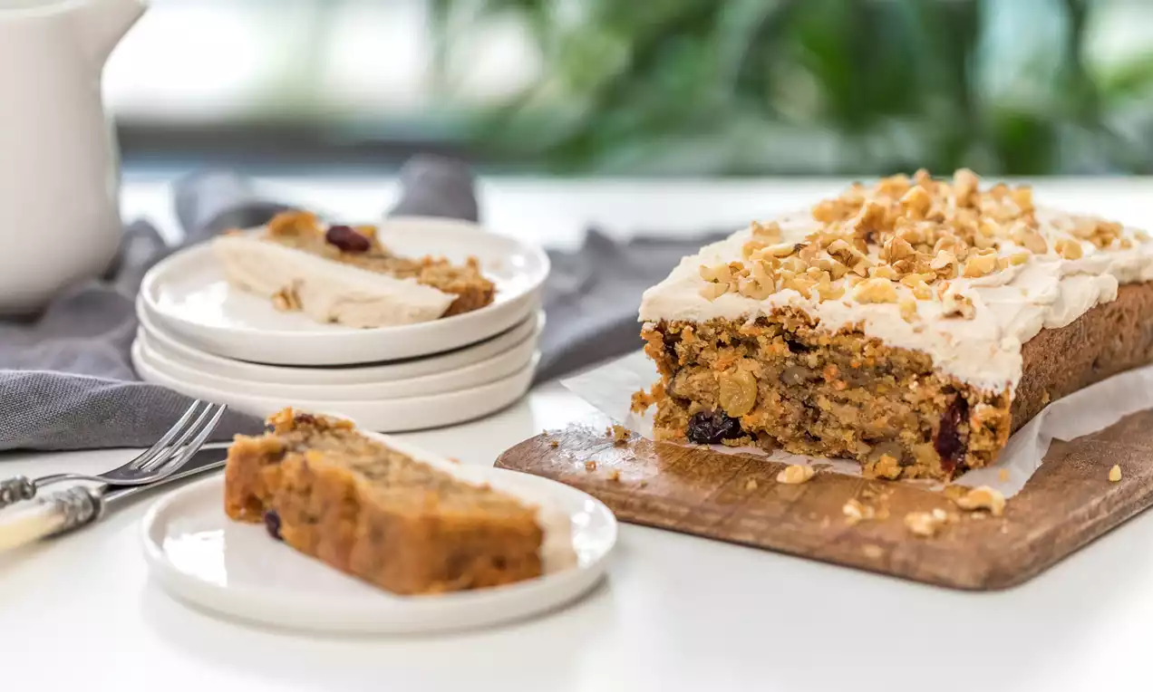 Bizcocho rectangular de zanahoria anaranjado con trozos visibles, cubierto con crema blanca y nueces, servido en un plato blanco