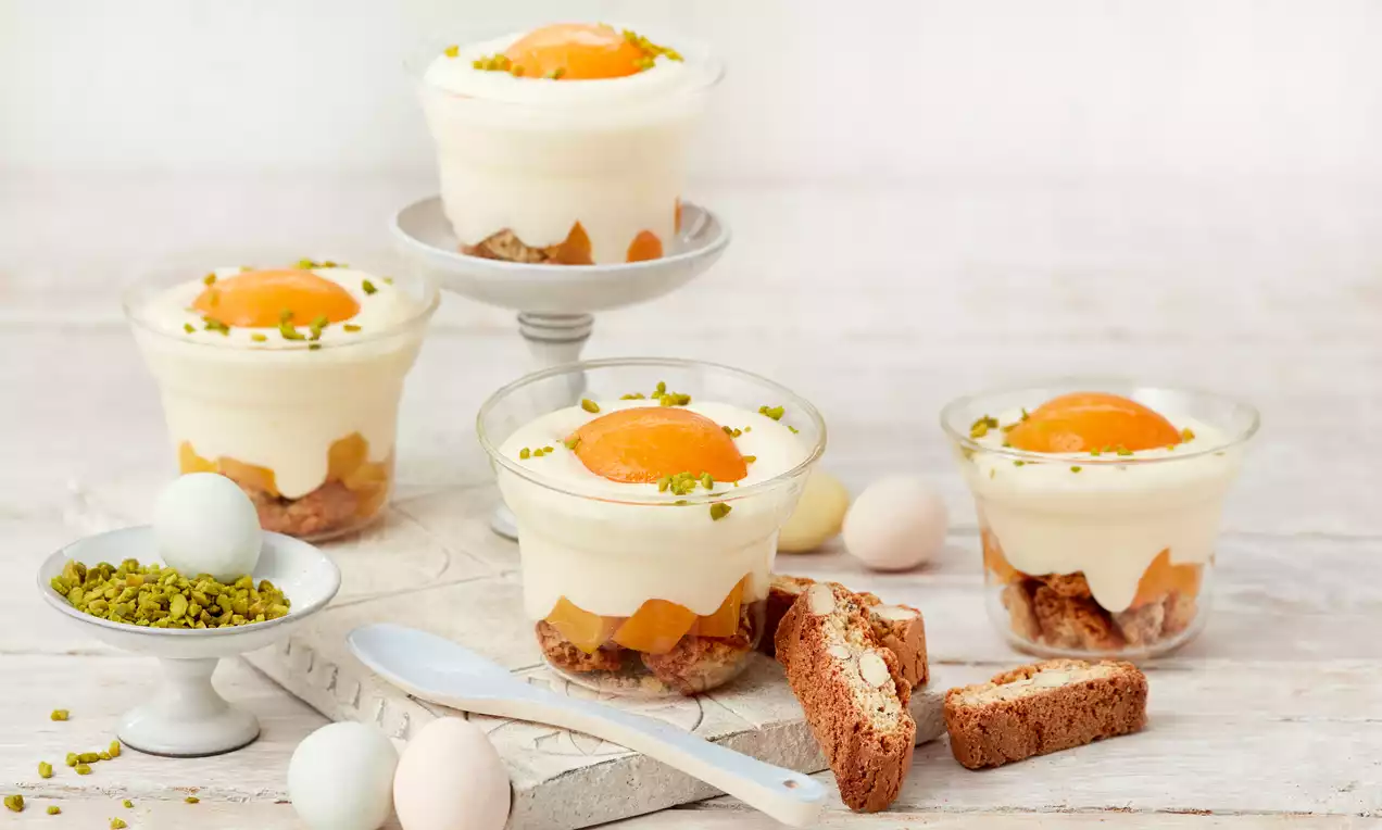 Dessert im Glas mit weißer Creme, orangefarbener Aprikosenhälfte, zerbröseltem Keksboden und Pistazienstreuseln auf hellem Holztablett