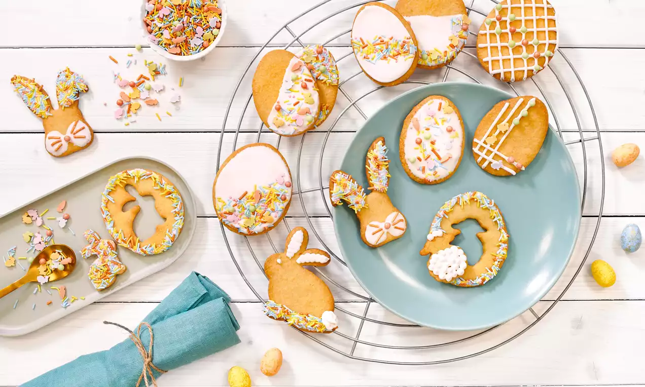 Golden brown Easter cookies shaped like bunnies and eggs, decorated with white icing and pastel sprinkles, arranged on plates and a cooling rack
