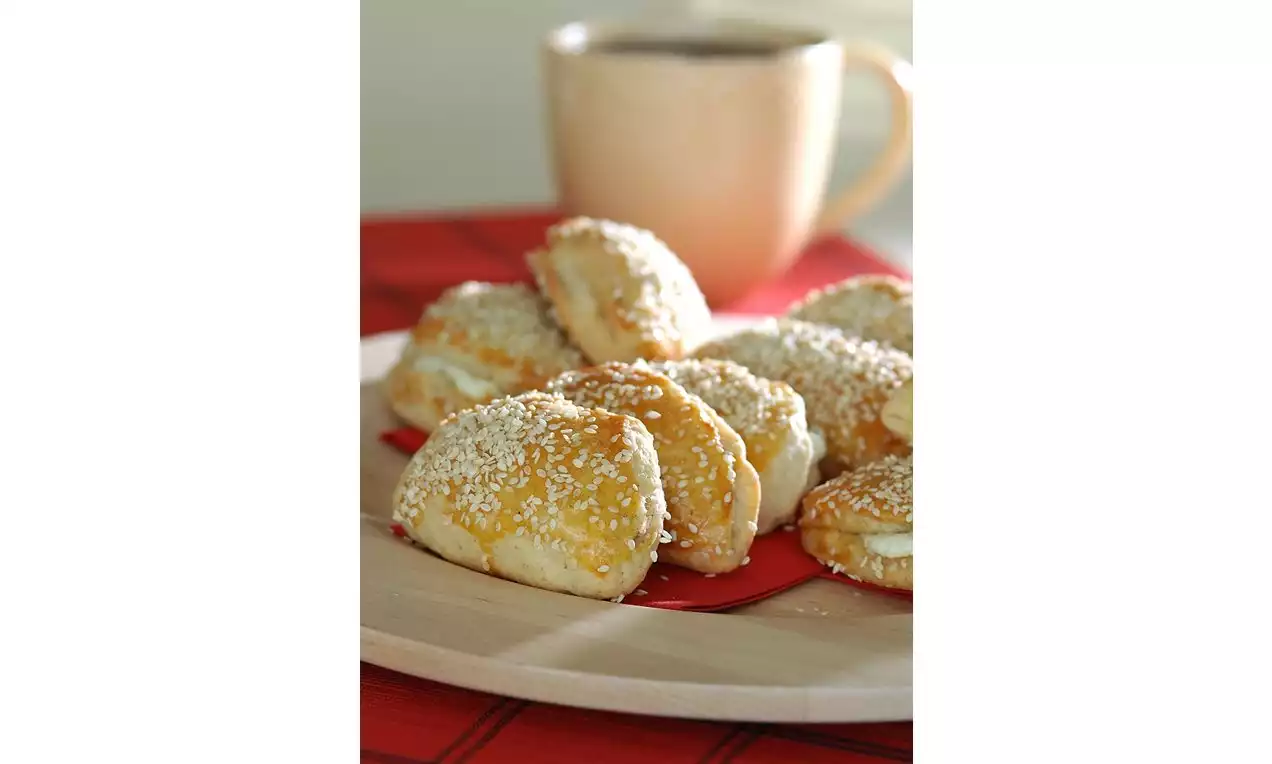 Altın rengi kepekli poğaçalar, üzerleri beyaz susamla kaplı, ahşap bir tabakta kırmızı bir peçete üzerinde sıcak içecekle sunulmuş