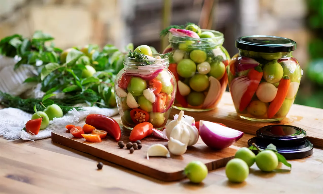 Светлозелени джанки, яркочервени чушлета и снежнобял чесън в ножично подредени буркани, поставени върху дървена маса с пресни подправки