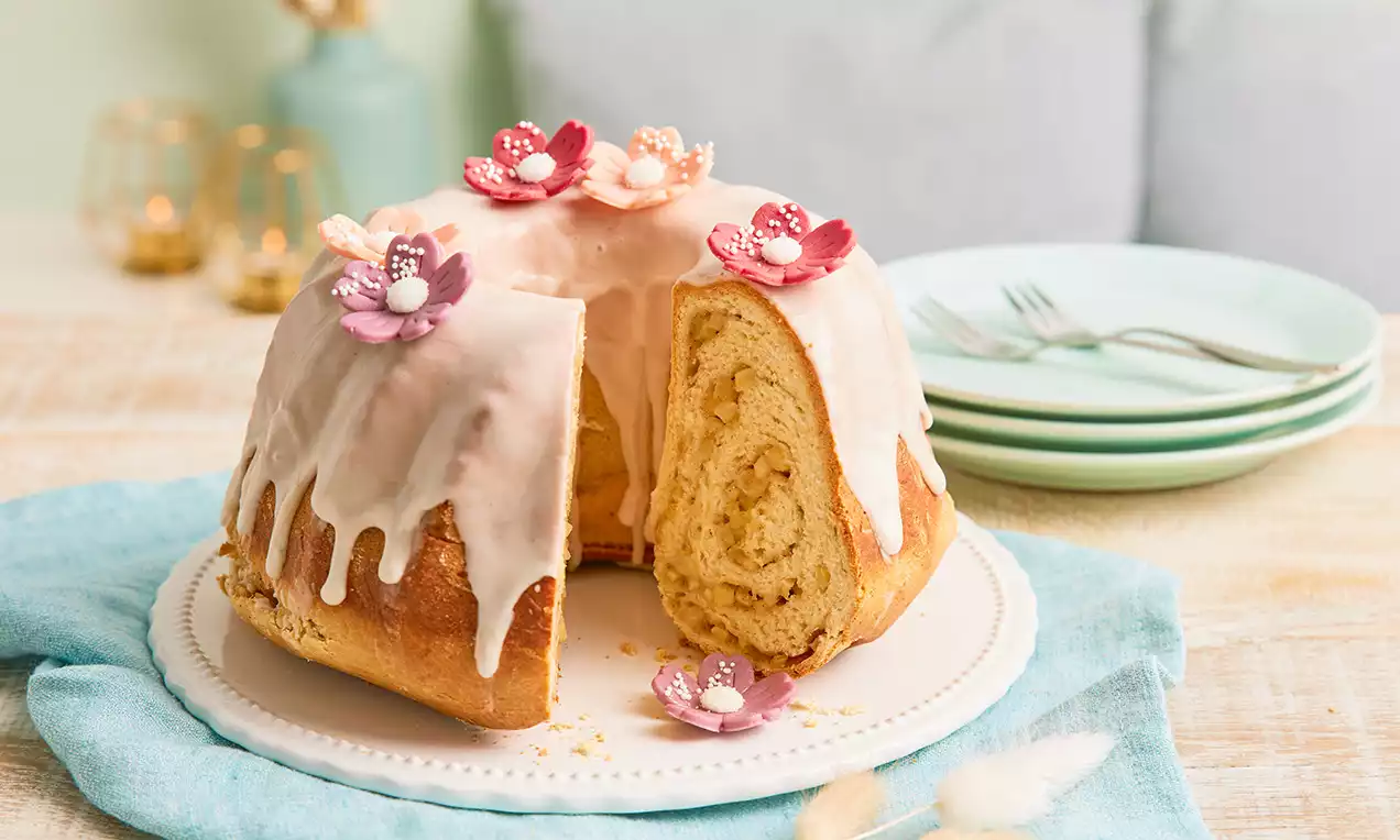 Apfel-Gugelhupf mit heller Glasur und filigranen Blüten dekoriert, auf weißer Platte mit türkisfarbenem Stofftuch angerichtet