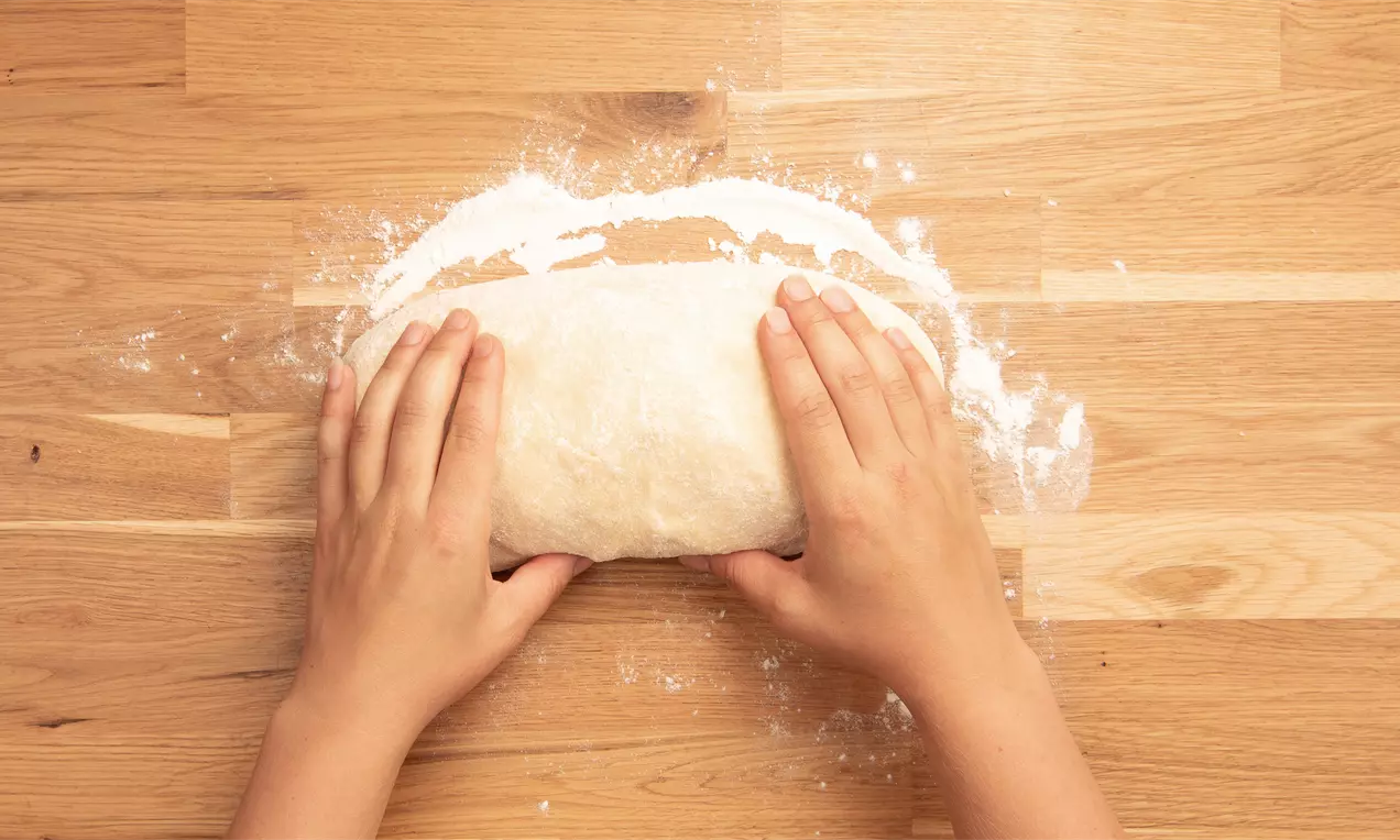 Ръце нежно оформят меко тесто с гладка текстура, поръсено с бяло брашно върху светъл дървен плот