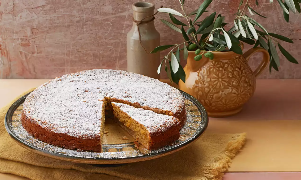 Runde Torte mit orangebrauner, saftiger Oberfläche, bestäubt mit Puderzucker, angeschnitten auf einer gemusterten Glasplatte auf gelbem Stoff