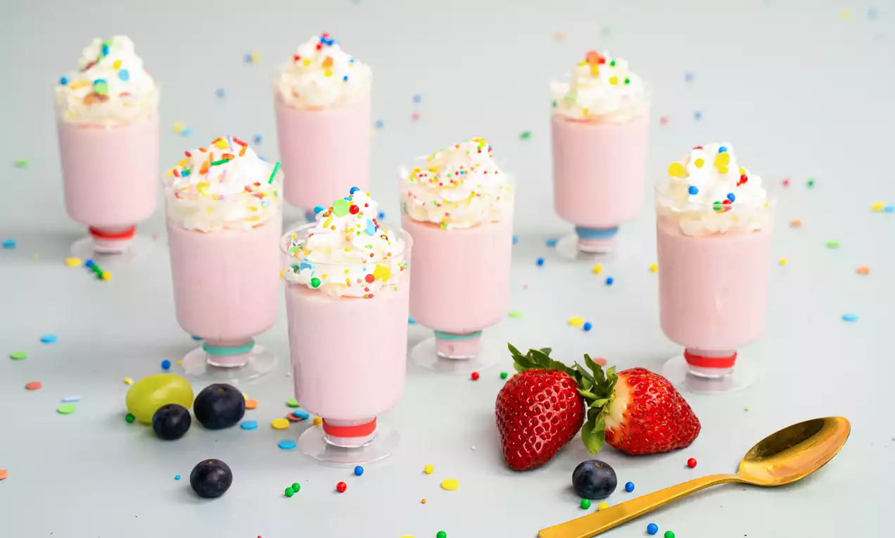 Roze kwarktaartglaasjes met toefjes slagroom en gekleurde sprinkels, omringd door aardbeien, bessen, een gouden lepel en confetti.