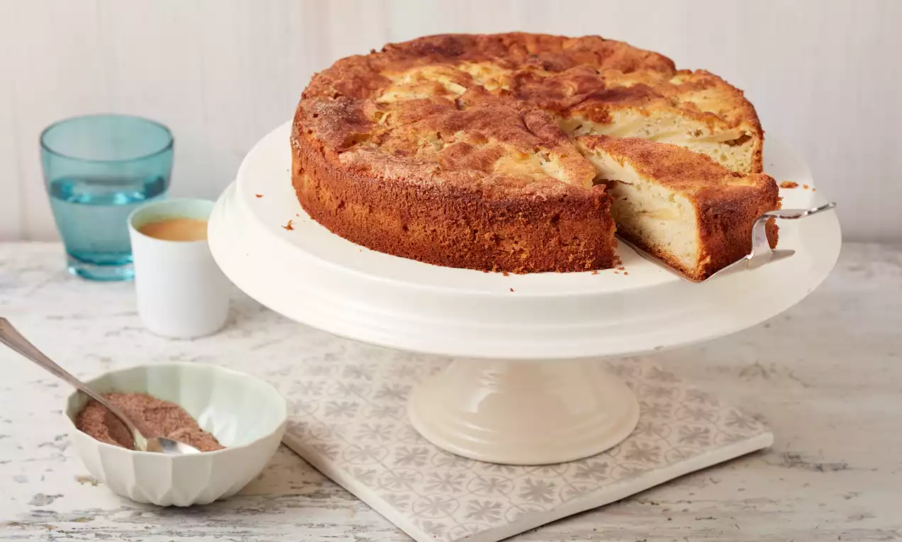 Apfelkuchen mit goldbrauner Kruste, saftigen Apfelstücken und leicht gebräunter Oberfläche auf weißer Tortenplatte mit angeschnittenem Stück
