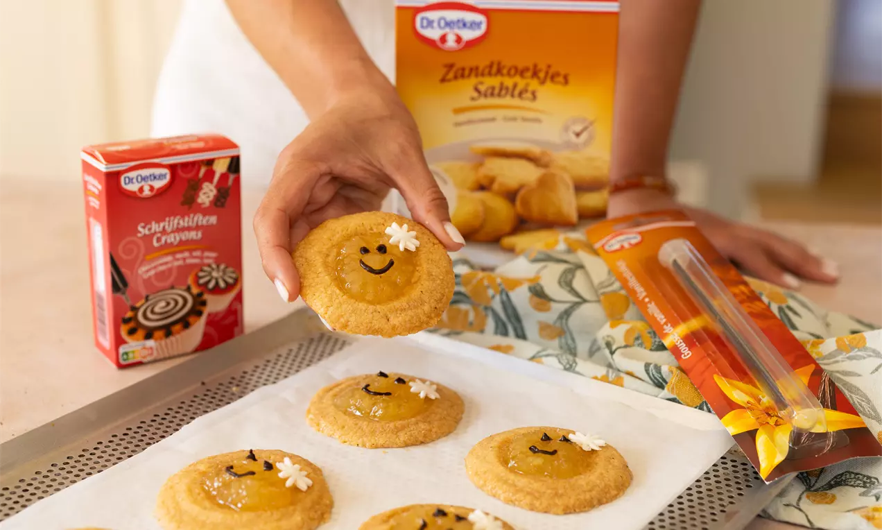 Goudbruine ronde koekjes met een glimlach van chocolade en witte bloemdecoratie op bakpapier, terwijl een hand er één omhoog houdt