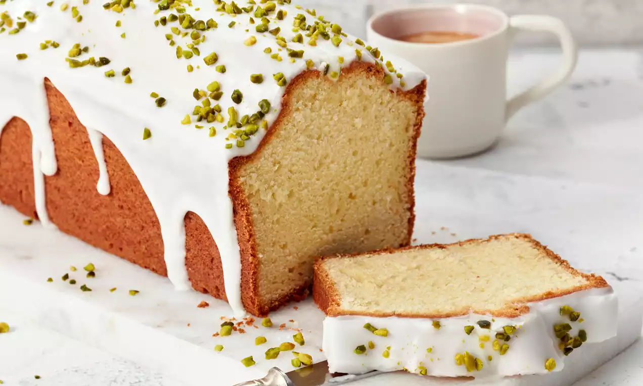Saftiger, goldbrauner Joghurtkuchen mit weißem Zuckerguss und gehackten Pistazien, serviert auf einer weißen Platte mit Kaffeetasse im Hintergrund